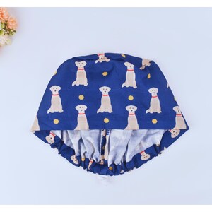 May include: A blue and white scrub hat with a pattern of yellow Labrador Retrievers. The hat has a gathered elastic band around the bottom.