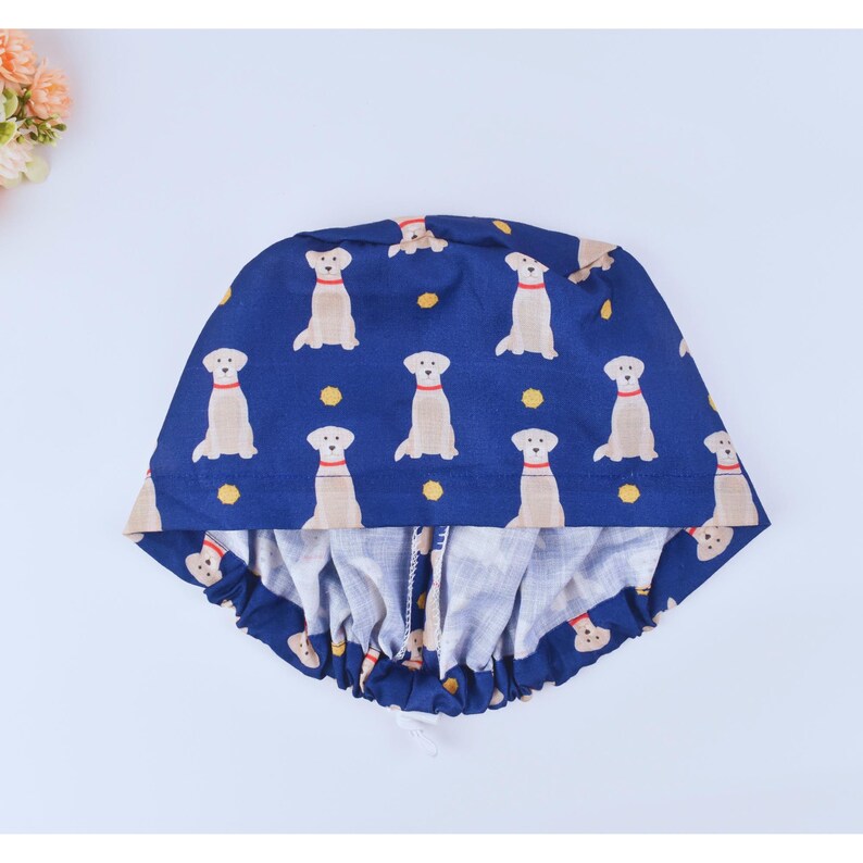 May include: A blue and white scrub hat with a pattern of yellow Labrador Retrievers. The hat has a gathered elastic band around the bottom.