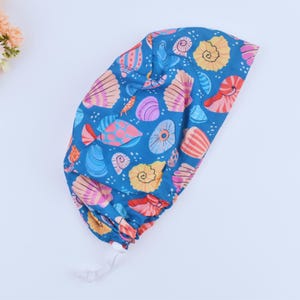 Peut inclure: Un bonnet de chirurgien bleu avec un motif de coquillages colorés. Le bonnet a un cordon de serrage blanc.
