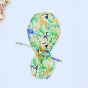 May include: A green, blue, and yellow patterned surgical scrub hat with a jungle animal theme. The hat features giraffes, zebras, snakes, toucans, and elephants.