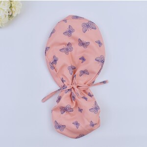 Puede incluir: Un gorro quirúrgico rosa con un patrón de mariposas azules. El gorro tiene lazos que se pueden atar en la parte posterior del cuello.