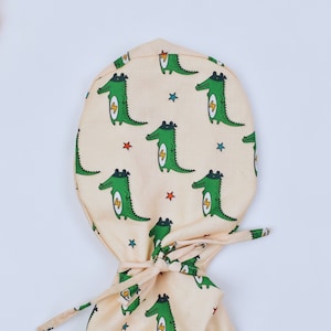 May include: A light beige scrub hat with a pattern of green crocodiles wearing superhero capes and lightning bolts. The hat has a tie at the back.