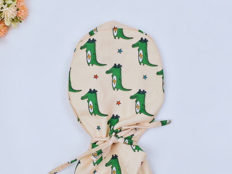 May include: A light beige scrub hat with a pattern of green crocodiles wearing superhero capes and lightning bolts. The hat has a tie at the back.