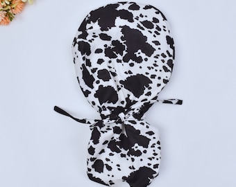 May include: A black and white cow print surgical scrub hat with a tie at the back.