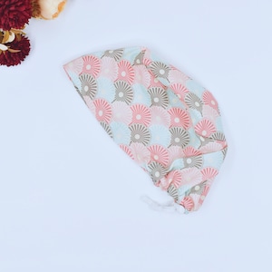 Op de afbeelding: Een patroon chirurgische scrub cap met een herhalend ventilatorontwerp in roze, grijs en lichtblauw. De cap heeft een wit trekkoord aan de achterkant. De achtergrond is wit.