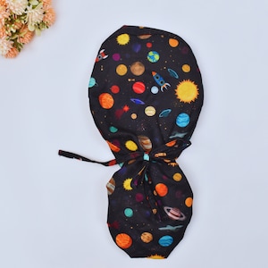 Op de afbeelding: Zwarte scrub cap met een kleurrijk ruimte-thema patroon. Het ontwerp bevat planeten, raketten, sterren en zonnen. De cap heeft een striksluiting voor een verstelbare pasvorm.