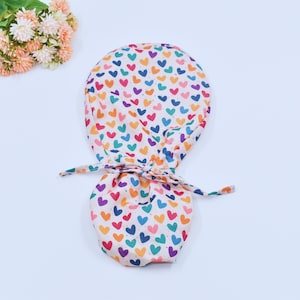 May include: A white scrub hat with a colorful heart pattern. The hat has a tie that can be adjusted to fit.