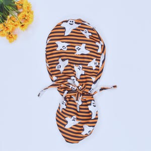 May include: A Halloween-themed scrub cap featuring a pattern of white ghosts on a background of orange and black horizontal stripes. The cap is tied at the back with matching fabric ties. The cap is made of a soft, flexible material.