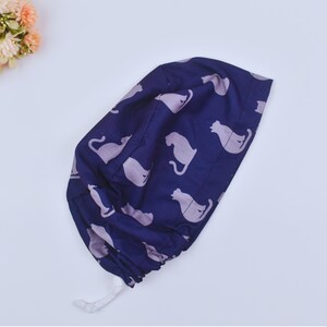 Puede incluir: Un gorro quirúrgico azul marino con un patrón repetido de siluetas de gatos grises claros. El gorro tiene un cierre de cordón y está hecho de una tela suave y flexible. El fondo es blanco.