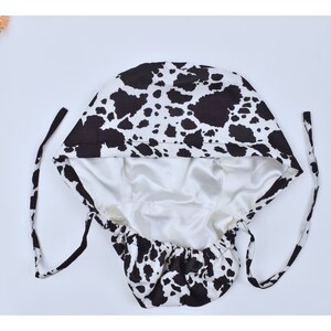 May include: A black and white cow print satin sleep cap with a white satin lining and adjustable ties.