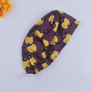 Puede incluir: Un gorro de quirófano morado con un patrón de lunas amarillas y murciélagos negros. El gorro tiene un cordón de ajuste blanco y un botón blanco.