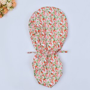 Op de afbeelding: Een witte stoffen muts met een rood en groen bloemenpatroon. De muts heeft een strik aan de achterkant.