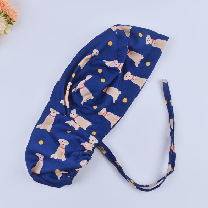 May include: A blue surgical cap with a pattern of yellow Labrador Retrievers wearing red collars. The cap has a drawstring tie.