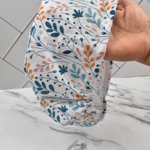 Puede incluir: Un gorro de quir&oacute;fano blanco con un patr&oacute;n floral en tonos azules, naranjas y rosas. El gorro tiene una banda el&aacute;stica fruncida en la parte inferior.