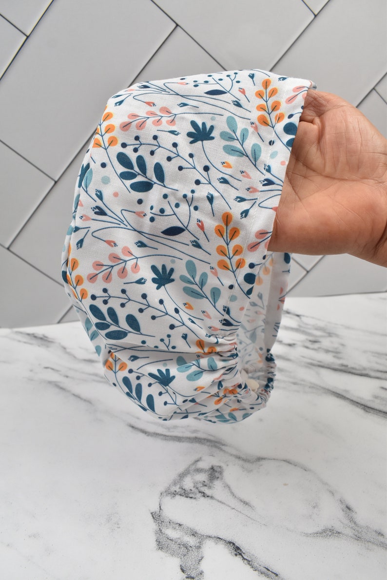 Puede incluir: Un gorro de quir&oacute;fano blanco con un patr&oacute;n floral en tonos azules, naranjas y rosas. El gorro tiene una banda el&aacute;stica fruncida en la parte inferior.