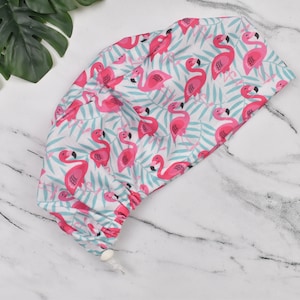 Op de afbeelding: Een witte scrub-muts met een roze flamingo-print. De muts heeft een trekkoord en een knoop aan de achterkant.