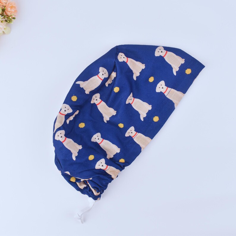 May include: A blue scrub hat with a pattern of yellow Labrador Retrievers. The hat has a drawstring.