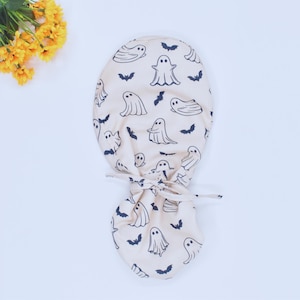 Puede incluir: Un gorro de quirófano beige con un patrón repetido de fantasmas y murciélagos negros. El gorro está atado en la parte posterior con una corbata de tela a juego. Se ven flores amarillas.