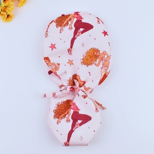 Könnte beinhalten: Rosa OP-Mütze mit Schleife. Die Mütze hat ein sich wiederholendes Muster von Frauen in rosa und roten Trainingsoutfits mit Sternen und anderen abstrakten Designs.