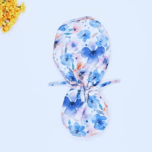 May include: A white scrub hat with a blue and pink watercolor floral print. The hat has ties that can be tied at the back of the head.
