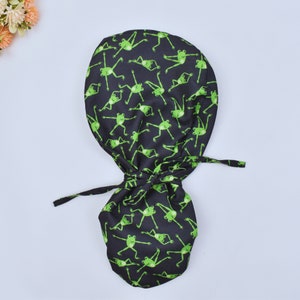 Op de afbeelding: Een zwarte scrubcap met een herhaald patroon van felgroene cartoonkikkers. De cap is aan de achterkant vastgebonden met lange stoffen banden. De kikkers staan in verschillende poses, wat een speelse touch aan het ontwerp geeft. De cap is waarschijnlijk gemaakt van een zachte, ademende stof.
