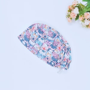 Puede incluir: Un gorro quirúrgico estampado con un diseño repetido de gatos de dibujos animados en varios colores. El gorro es principalmente rosa, azul y blanco, con un cierre de cordón. El fondo es blanco.