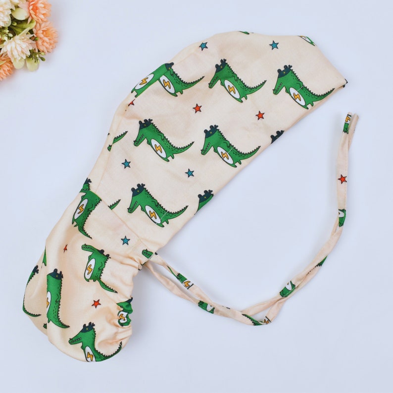May include: A beige surgical cap with a pattern of green crocodiles with yellow lightning bolts and red stars. The cap has a long tie that can be tied at the back of the neck.