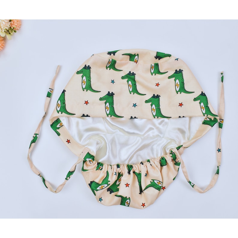 May include: A beige satin sleep cap with a green alligator print and stars. The cap has a drawstring closure.