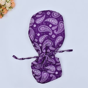 Op de afbeelding: Een paarse bandana met een wit paisley-patroon. De bandana is bovenaan vastgebonden en de uiteinden hangen naar beneden.