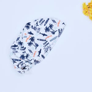 May include: White fabric hat with a blue and orange print of palm trees, surfboards, and sharks. The text "Surfing", "Waves", and "Surfing" is printed on the hat.