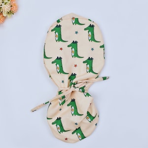 May include: A beige fabric scrub hat with a drawstring closure. The hat features a repeating pattern of green cartoon crocodiles with lightning bolts and stars.