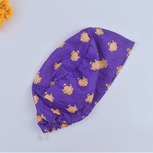 May include: A purple surgical scrub cap with a repeating pattern of tan ghosts and bones. The cap is gathered at the back and has a white drawstring. The background is a light purple.