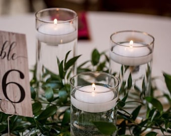 Pode incluir: Três cilindros de vidro transparente com velas flutuantes e vegetação sobre uma toalha de mesa branca. As velas são brancas e a vegetação é verde. O número da mesa "6" é visível em primeiro plano.