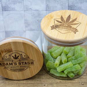 May include: Two clear glass jars with bamboo lids. One jar is open, filled with green, sugared sweets. The lids are engraved with cannabis leaf designs and the text "ADAM'S STASH" and "Cannabis KATEY'S STASH".