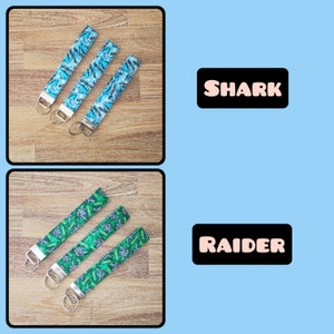May include: Three blue keychains with a shark print design and three green keychains with a raider print design. The keychains are on a wooden surface.