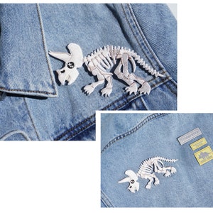 May include: Two denim jackets with white embroidered dinosaur skeletons. One jacket has a small label that says "Triceratops" and a smaller label with a dinosaur silhouette. The other jacket has a larger dinosaur skeleton patch.