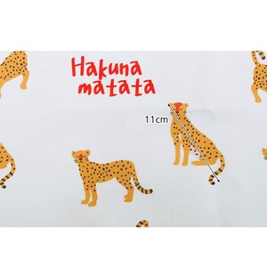 May include: White fabric with a repeating pattern of orange and black cheetahs. The text "Hakuna Matata" is printed in red letters. The fabric is 11 inches wide.
