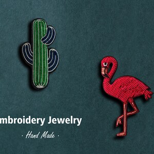 May include: Two embroidered jewelry pieces: a green cactus and a pink flamingo. The text "Embroidery Jewelry. Hand Made." is below the pieces.