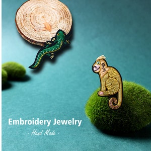 Puede incluir: Dos broches bordados, uno de un lagarto verde y otro de un mono amarillo, se muestran sobre un fondo verde. El texto "Embroidery Jewelry - Hand Made -" es visible debajo de los broches.