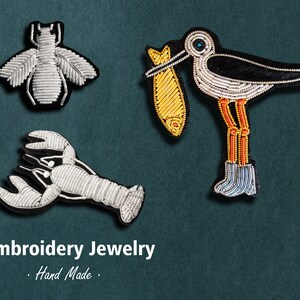 May include: Three embroidered patches featuring a bee, a lobster, and a bird carrying a fish. The patches are silver, black, and gold. The text "Embroidery Jewelry. Hand Made." is below the patches.