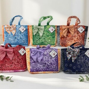 May include: Six rectangular tote bags featuring paisley bandana patterns in blue, green, orange, red, purple, and dark blue. Each bag includes two top handles, a decorative fabric bow on the front, and a small white rectangular tag attached to the side.