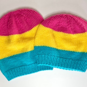 May include: Two knitted beanies in the colors of the Pansexual Pride flag: pink, yellow, and turquoise. The hats are made of a soft, textured yarn and are likely handmade. The beanies are a comfortable, stylish accessory.