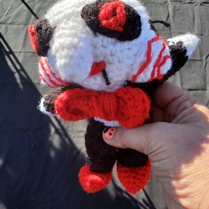 May include: A crocheted plush toy with a white body, black and red striped arms, red bow tie, black legs, and red shoes. The toy has a black and red eye.