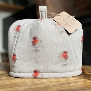 Könnte beinhalten: Ein beiger Teewärmer mit einem sich wiederholenden Muster von Aquarell-Rotkehlchen mit roten Brüsten. Der Teewärmer hat einen Schlaufengriff und ein Etikett mit dem Text "Handmade with love".