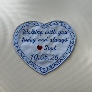 Può includere: Una toppa a forma di cuore con bordatura ricamata blu e testo. Il testo recita "Walking with you today and always Dad 10.05.26" con un simbolo a cuore rosso. La toppa è bianca con ricami blu.