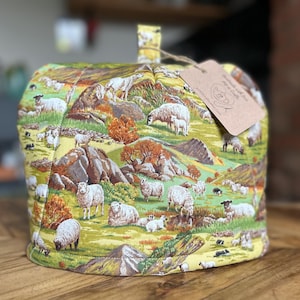 以下が含まれることがあります： 緑、黄色、茶色の牧歌的な羊のシーンが描かれたキルトのティーコージー。コージーにはループハンドルとタグが付いています。生地は、木や岩のある丘陵地帯で草を食べる羊を描いています。