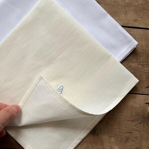 May include: Two white linen tea towels, one with a blue embroidered design. The towels are folded and stacked on a wooden surface.