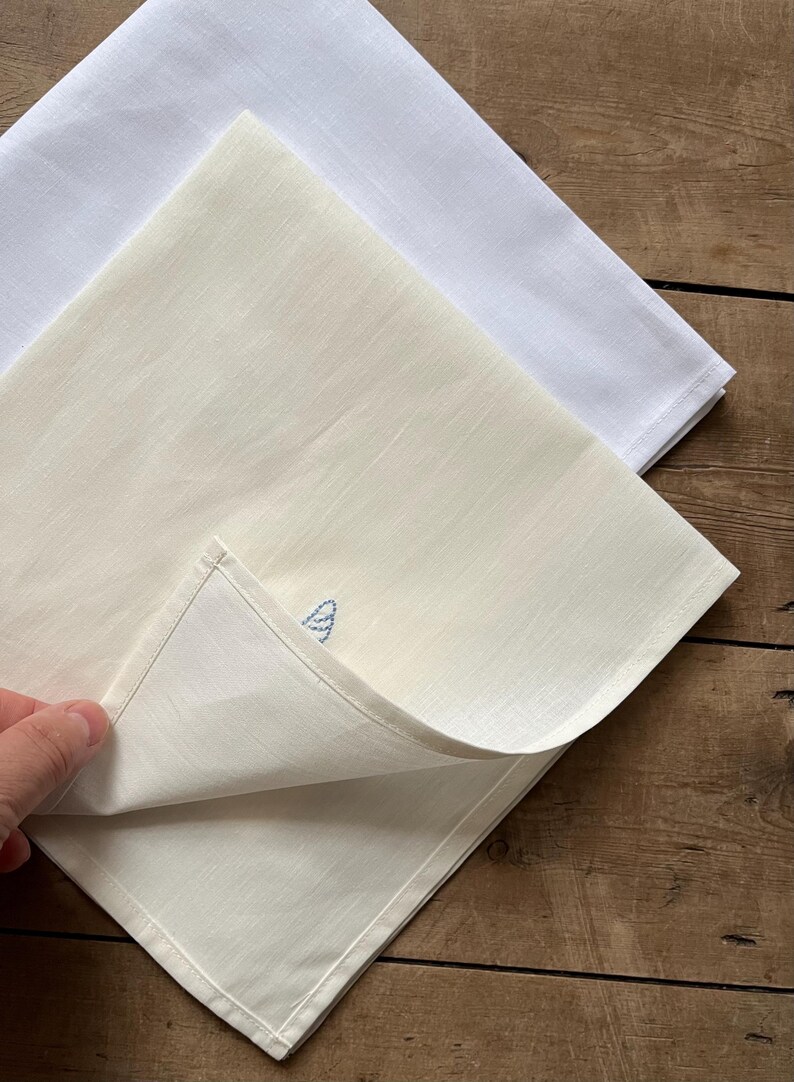 May include: Two white linen tea towels, one with a blue embroidered design. The towels are folded and stacked on a wooden surface.