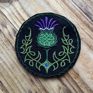 Op de afbeelding: Een zwarte geborduurde patch met een groen en paars distelontwerp. De distel is omgeven door een groen Keltisch knoopontwerp.