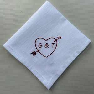 May include: A square white handkerchief featuring a dark red embroidered design of a heart pierced by an arrow. The letters G & T are stitched in dark red thread inside the centre of the heart. The fabric has a visible hemmed border.